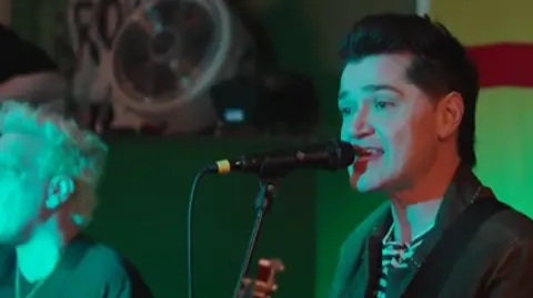 A man with short dark hair and a dark jacket in front of a microphone in a room with green lighting