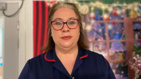 Emma Stewart is wearing a red and blue blouse and red glasses. She is standing in front of a Christmas themed background inside the hospital 