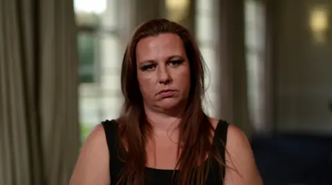 A woman wearing a black tank top and long reddish brown hair sits and stares at the camera