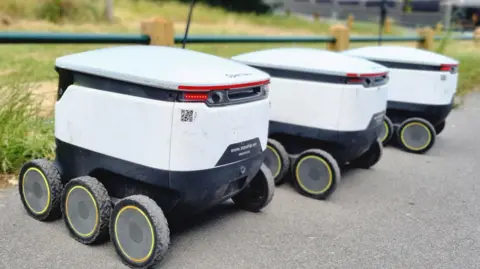Three white robots with black wheels parked up alongside each other on tarmac.
