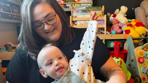 Emma Baugh/BBC Wren wearing a green pinafore and white shirt with black dots sitting in Naomi's lap - wearing  black shirt 