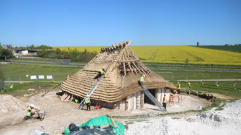 English Heritage A replica neolithic hall under construction near Stonehenge with multiple volunteers in high-vis working on the structure.