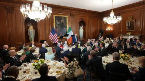 PA Media People sitting around round tables in a room. There is wood panelling on the walls and two chandeliers hanging from the ceiling. Donald Trump is addressing the crowd and an American and Irish flag are behind him.
