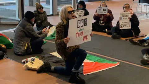 Protesters insid BBC Scotland HQ reception. There is a woman at the front kneeling down holding a protest sign with a Palestinian flag lying on the floor next to her. There are five other people sitting on the ground around her also holding protest signs.