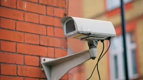 A CCTV camera fixed to a wall on a street corner.