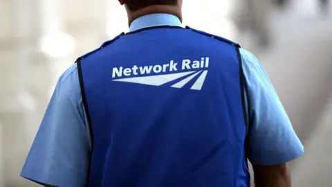 A generic shot of a Network Rail worker from behind. They are wearing a blue vest with the company branding on the back. 