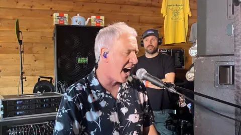 Clint Boon of Inspiral Carpets sings into a microphone. He is surrounded by sound equipment and a man wearing headphones and a baseball cap is stood behind him. 