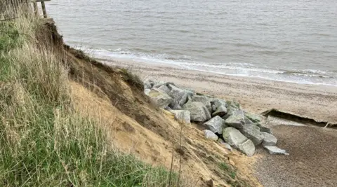 GUY CAMPBELL/BBC Grey, granite boulders can be seen at the base of a steep, sandy cliff with a grassy area in the foreground and the sea in the background