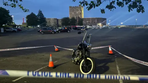 A black Harley-Davidson motorbike at a car park sectioned off with cones and police tape. It is night, a castle can be seen in the background and some street lights.