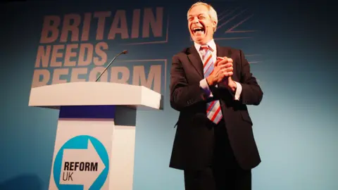 PA Media Reform UK leader Nigel Farage stands on stage in Cornwall at a rally next to a white podium with the party symbol on it. He is smiling with his mouth wide open and is stood in front of a backboard which says "Britain Needs Reform".