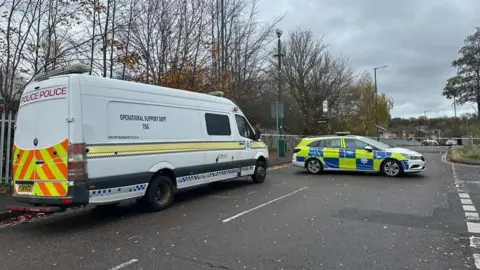 A large white police van parked next to a police car on a two-lane road. A cordon can be seen horizontally at the back of the image.
