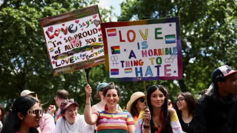Reuters A crowd of smiling attendees at a Pride event under a canopy of green trees. In the centre, two women hold up colorful, hand-painted protest signs; the sign on the left reads "Love says I'm so proud of all of you," while the larger sign on the right reads "Love is a horrible thing to hate" alongside illustrations of various Pride flags.