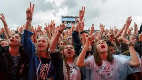 EPA A group of fans at the front of a crowd at an outdoor music event. They are all young people. They have their arms raised and are looking towards the stage.