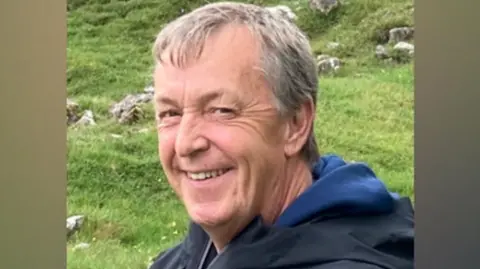 Shaun Flatley, who has short, grey hair, looks at the camera. He is wearing a blue hoodie with a dark navy coat over it. Grass and rocks are pictured behind him. 