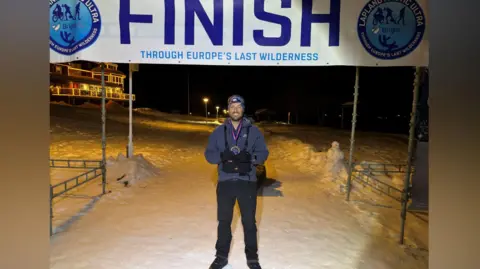 Rob Wilkinson A picture of Rob Wilkinson at the end of the Lapland Arctic Ultra challenge. Rob, who is wearing a blue jacket and black trousers, has his medal around his neck. The ground is covered in snow and a banner indicating the finish line can be seen above his head.
