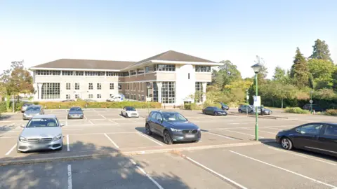 Cars are parked in marked bays in front of a large two‑storey office building in Surrey, with vehicles and trees in the background under a blue sky.