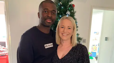 Alexis Brown Man in black jumper which says 'religion' on a white background with a blonde woman in a sparkly dress. They are both looking at the camera and there is a Christmas tree behind.