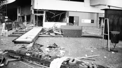 The remains of a pub after a bombing, showing blown out windows on the ground floor, hanging rafters and debris spilling into the street. An external wall sign saying Brew is just visible on a corner leading away from the main door. The pub's main sign lies on the floor in the foreground, broken so only some of the letters can be seen.