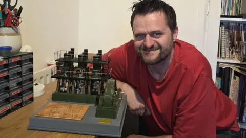 Lee Smithson Lee sits at a wooden desk in a well-organised workspace. On the desk, there is a detailed scale model of an industrial machine or factory setup, featuring green structures and intricate components. To the left of the desk, there is a stack of small labeled drawers, likely used for storing tools or model parts, along with a red spray can and a pair of scissors in a holder. Behind him, there is a white wall with framed artwork and photographs hanging on it. On the right side, there is a bookshelf filled with books and magazines, and a wooden chair is partially visible in front of it.