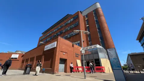 Stuart Woodward/BBC Redstone House pictured on a sunny day. It is a large brick building, several levels tall with windows lining each floor. A few people are gathered outside it.