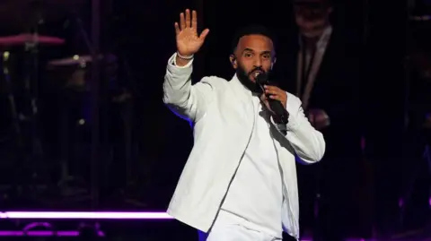PA Media The singer is wearing a white suit with white long-sleeved top beneath. He is holding a microphone to his mouth in the image