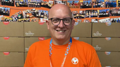 Rob Wallis wearing an Orange T Shirt stood in front of stacked kindness boxes.