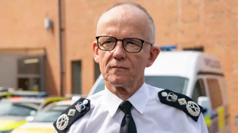 Sir Mark Rowley is seen in uniform standing in a police station car park with police vehicles visible behind him
