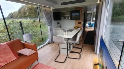 Capsule Holiday Homes/Alice Gregory A bright, modern interior of a compact modular home with floor-to-ceiling windows. The space includes a small kitchen with a white island and bar stools, light wood flooring, and minimalist cabinetry. 