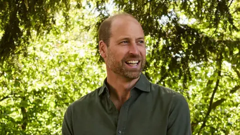 Prince William smiles broadly as he stands in front of trees during an event. He is wearing a dark green shirt.
