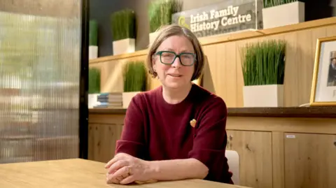 EPIC Fiona Fitzsimons, a woman with short, blonde hair cut in a bob, looks at the camera while sitting in an office in the Irish Family History Centre. She is wearing a pair of green, square-framed glasses and a claret-coloured crew-neck jumper. She is seated at a wooden table and there wooden cupboards and an opaque glass screen behind her. 