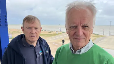 Mark Rose and Nigel Sutton posing for the camera. A beach and the sea are visible behind them.