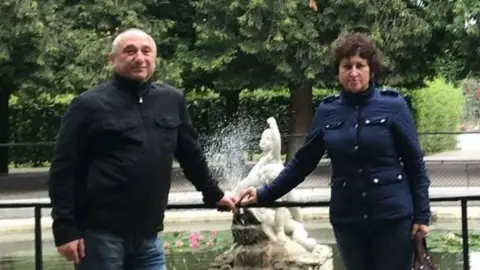 GoFundMe A man in a zipped-up black jacket and a woman in a navy blue jacket with buttons pose for a picture in front of a fountain in a park. Each has an arm outstretched with their fingers touching each other's.
