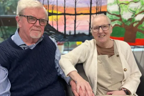 Sally and Kevin are looking at the camera, holding hands and smiling. Kevin is wearing a blue and white stripped shirt and a navy knitted tank top. Sally is wearing a cream cardigan, brown t-shirt and light brown dungarees. Both have short white hair and glasses.