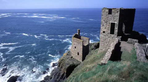 BBC This picture shows the iconic engine houses of the historic Botallack Mine on the rugged Cornish coastline. The structures are perched dramatically on cliffs overlooking the Atlantic Ocean, with waves crashing against the rocks below. These stone buildings were part of Cornwall’s tin and copper mining industry during the 19th century and are now preserved as part of the UNESCO World Heritage Site for Cornish Mining. The scene captures the wild beauty of the coast, with deep blue sea, clear skies, and the stark remains of industrial heritage blending into the natural landscape.