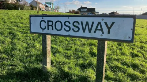 A close-up of a white road sign that reads CROSSWAY in black letters that is set in a grassy area with houses behind it.