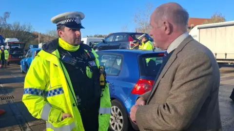 Suffolk Constabulary Sgt Alex Kelly (left) and Tim Passmore talking to each other at a check stop. Sgt Kelly is wearing a police hat, a high-vis jacket, and a black police uniform with cameras on his body. Tim Passmore is wearing a tweed jacket, with a shirt and tie. He has short fair hair. There are cars behind them and other police officers in the distance. 