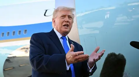 Trump wearing a suit and blue tie gesturing with his hands in front of microphones
