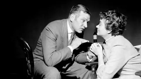 PA Media A black and white photo of a man and a woman. The man is wearing a suit and sitting on a chair holding the woman's hand. The woman has her eyes closed.