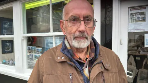 Carla Fowler/BBC John Freeman, a man with a white beard and glasses wearing a brown jacket standing in front of a shop window in Whitby