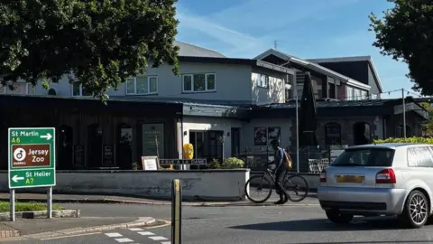 A picture of the Five Oaks area with the pub in the background. There is cyclist and a car going past.