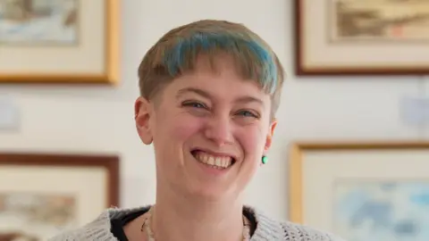 Jamie Niblock/BBC A woman with short cut strawberry blonde hair and dyed blue parts in a fringe. She is smiling and has one green earring. She is wearing a white knitted jumper. Pieces of framed artwork rest on the wall behind her.