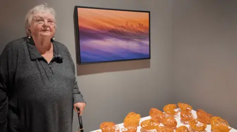 Patricia Tutt is standing by a photograph that she has taken of Peel Castle. The ocean is a blending shade of purple and blue, and the sky is a deep orange. 