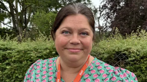 Luke Deal/BBC A woman stands outside on a grey day. She is smiling at the camera. She has dark hair that is tied back and wears a green dress with small pink roses over it. There is an orange lanyard around her neck.