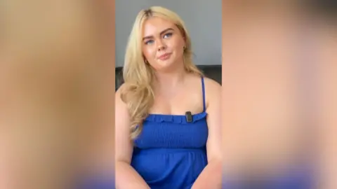 A girl with blonde hair is sitting with her head tilted to one side. She is wearing a blue summer dress.