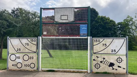 The concrete pitch in Hailey Park with the metal fence structures on one side. A basketball board can be seen as well as white metal sheets on either side which have circles with points assigned to them and an illustration of a football player diving. Trees can be seen in the background.