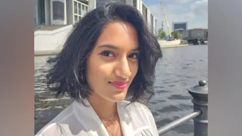Dr Syamala Roberts A 'selfie' of a woman wearing a white blouse. She has short brown hair and she is smiling.