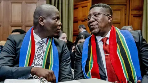 EPA Politisi Afrika Selatan Ronald Lamola dan pejabat lain - baik dalam pakaian pintar maupun dengan syal Afrika Selatan di leher mereka - berbicara ketika mereka duduk sebelum sidang di Pengadilan Internasional (ICJ) di Hauge - Januari 2024