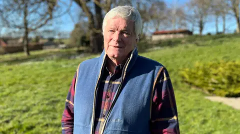 Alan Muir stands in the middle of the picture with a background of grass and trees. He is wearing a blue gilet over a red and navy chequered shirt. 