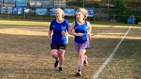Two women wearing blue tops, shorts and trainers, running on a rugby pitch