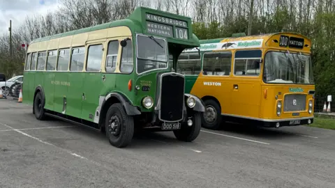 Two vintage buses parked in a car park. One has green and cream paintwork and a sign reading "Kingsbridge, Western National" on it, while the other is mustard yellow and green and says "Yatton" on the front. 
